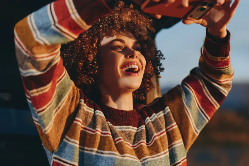 Cheerful woman outdoors, curly hair, wearing plaid sweater, smiling and laughing, arms raised in carefree lifestyle, warm sunlight, soft breeze, evening sky, natural golden colors