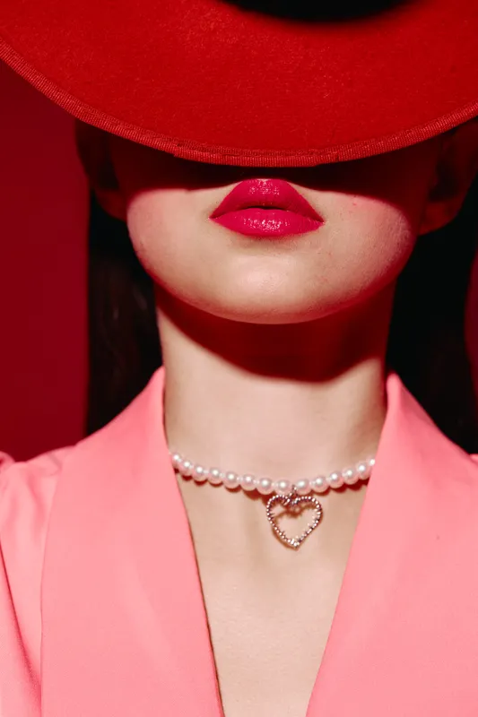 Close-up of a fashionable woman wearing bright red lips, a pearl necklace with a heart pendant, and a red hat shadowing her eyes. Creative beauty and fashion concept.