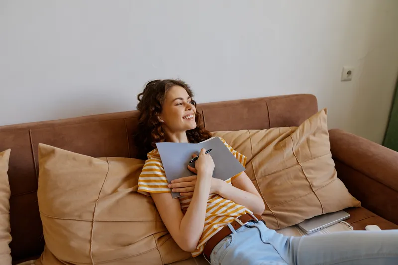 Young woman lounging on a brown sofa at home, smiling with a notebook in her hands, casual striped tee and jeans, warm light creating a relaxed lifestyle moment