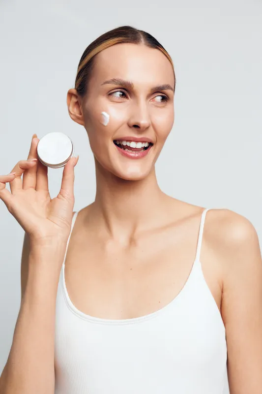 Young woman with clear skin applies cream on face holds cosmetic jar smiles looks aside isolated on light background skincare routine beauty concept natural makeup.