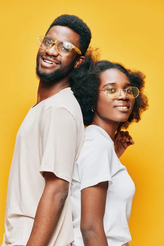 Two smiling models with glasses in casual outfits against a vibrant yellow background, showcasing a youthful and positive vibe
