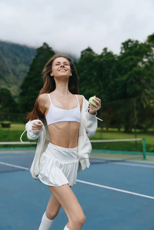 Young female tennis player enjoying a game, wearing a white sports outfit, evoking joy and confidence, set against a lush green background on a cloudy day
