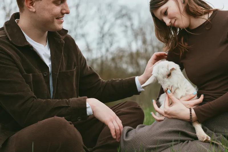 Goat kid cozy moment with a smiling couple outdoors, gentle interaction, sunlit meadow, casual clothing, warm colors, natural setting, quiet affection, wholesome lifestyle scene