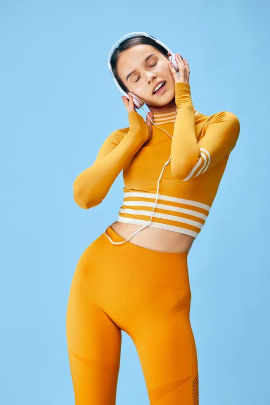 Fitness woman in vibrant orange athletic wear with headphones, posing in a studio with dynamic movement, conveying energy, focus and music-inspired performance in a bold sport style.