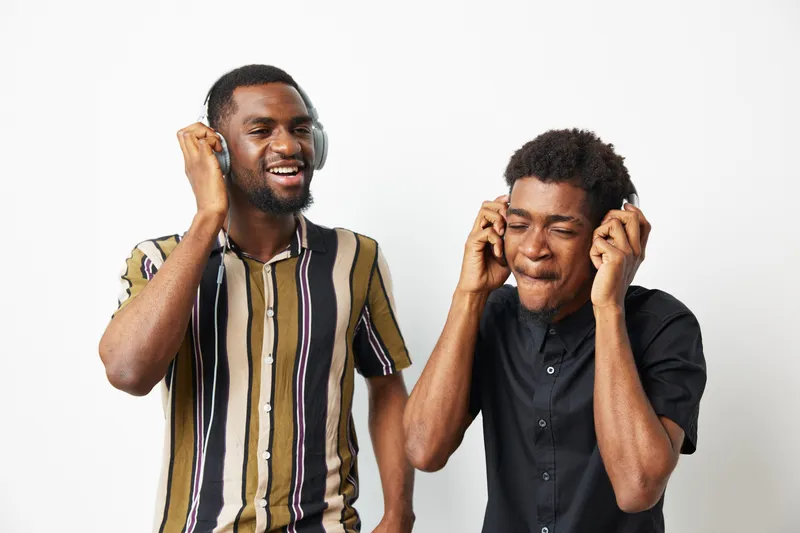 Two black men enjoying music together, wearing headphones. They express happiness and relaxation in a bright, minimalist setting. Perfect for capturing joy and friendship.