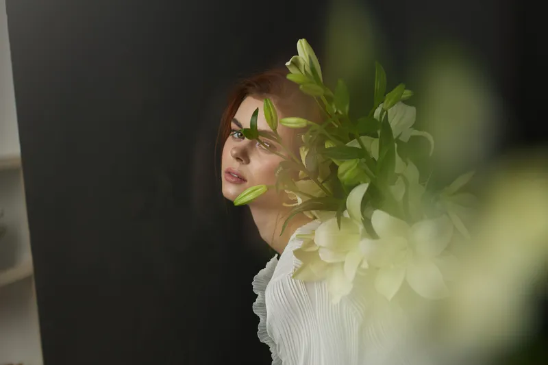 Woman with long red hair and white lilies against a dark background, expressing beauty and tranquility in a soft, seasonal floral arrangement