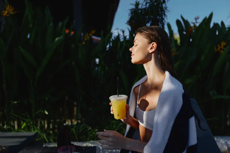 Woman outdoors, sipping juice, sunlit moment, relaxed posture, towel over shoulders, warm light, lush greenery background, casual summer vibe, candid lifestyle scene, bright colors, peaceful mood