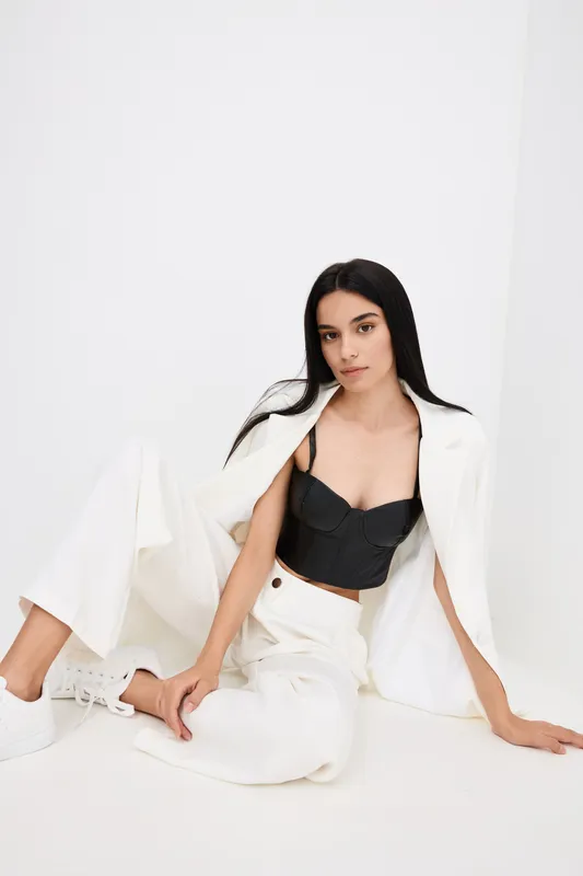young woman in white pantsuit and black corset top sitting on floor against white background with long black hair, casual elegance, modern fashion style