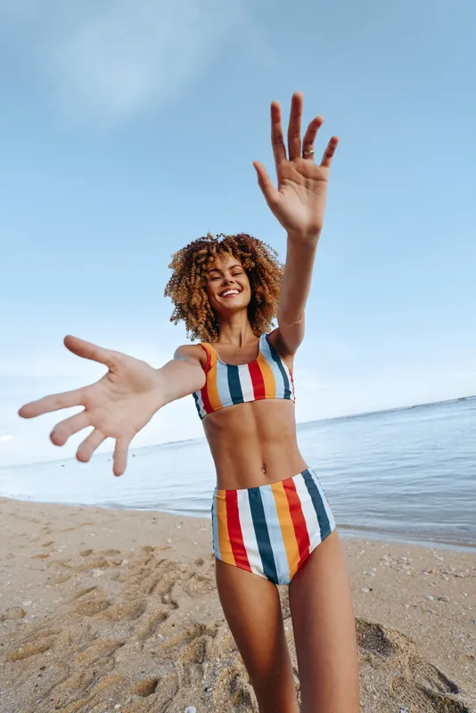 Woman smiling with curly hair on the beach, striped swimsuit, playful hand gesture, ocean water and blue sky behind, summer outdoors, carefree vacation mood