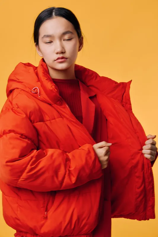 Young woman with long hair wearing bright orange puffer jacket over red sweater posing against yellow background. Fashion model showing winter clothing with closed eyes and soft expression.