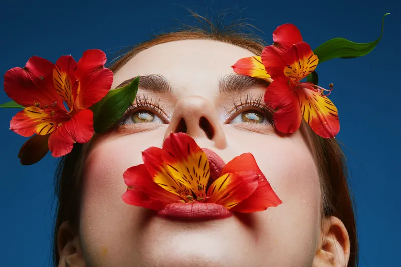 Close up portrait of a young woman with a serene expression, adorned with vibrant red and yellow flowers, against a deep blue background, highlighting beauty and nature