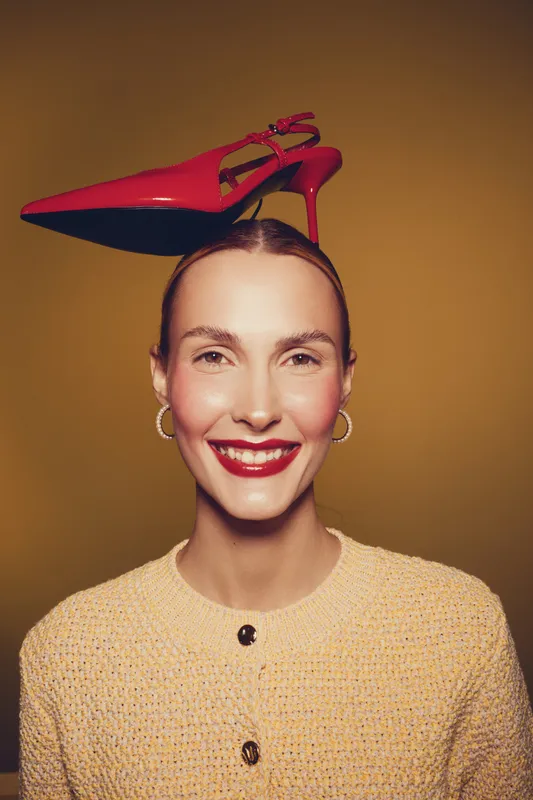 Smiling woman with red high heel shoe balanced on her head wears yellow textured sweater and gold hoop earrings. Studio portrait with warm brown background and playful fashion concept.