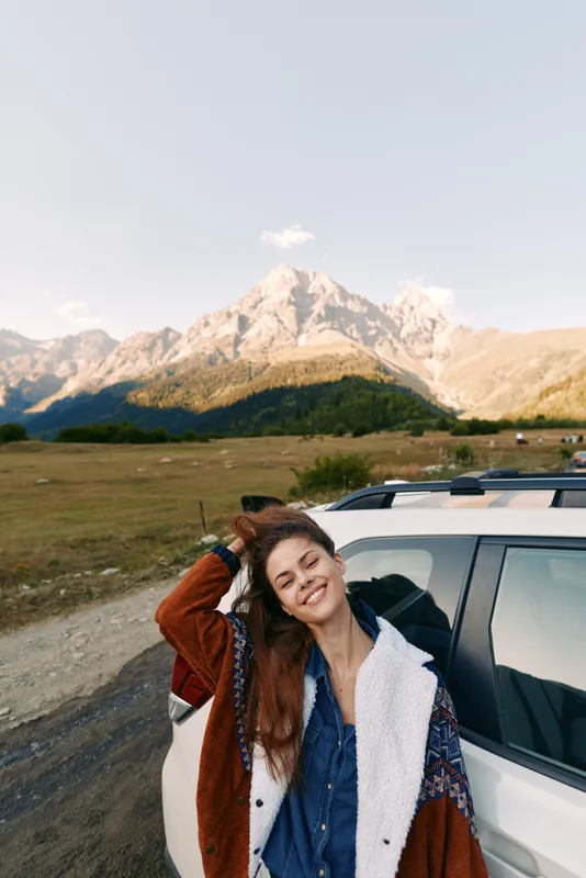 Woman leaning on car smiling beside mountain landscape during travel roadtrip, casual portrait in jacket, scenic outdoor adventure by vehicle on a rural road at golden hour.