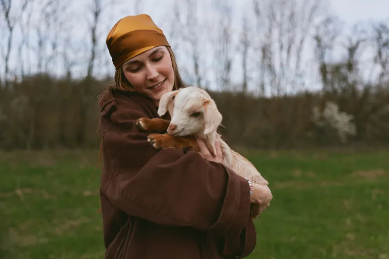 Young woman wearing a brown jacket and headscarf gently holds a baby goat outdoors in a green field during early spring, expressing warmth and connection with the animal.