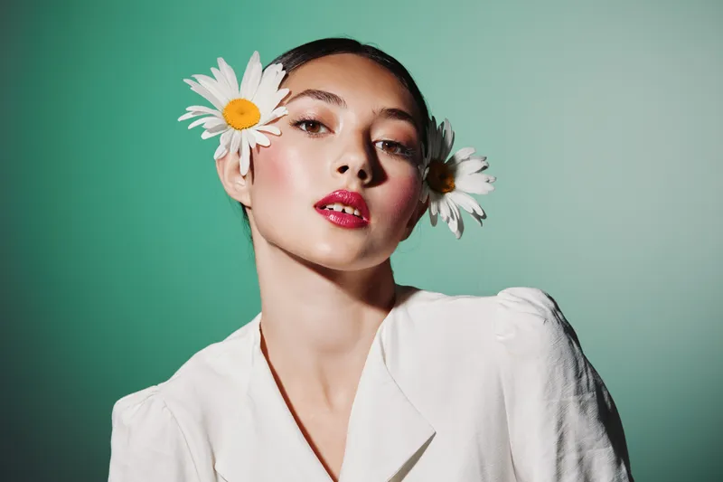Young woman with radiant makeup and glossy red lips wearing white with daisies in her hair, photographed on a mint green background. Concept for beauty, fashion, and creativity.