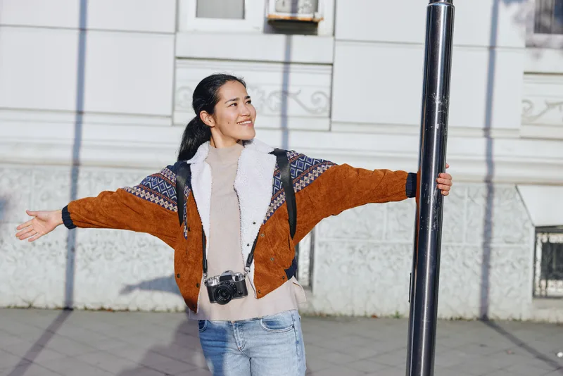 A woman with arms spread wide smiles while standing outdoors in a warm jacket and sweater on a sunny day, urban backdrop highlighting casual fashion and freedom