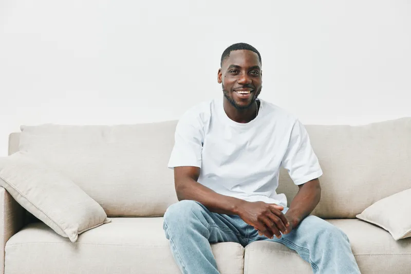 Smiling African American man wearing a white t shirt and jeans, sitting on a light couch, conveying happiness and relaxation in a minimalist setting