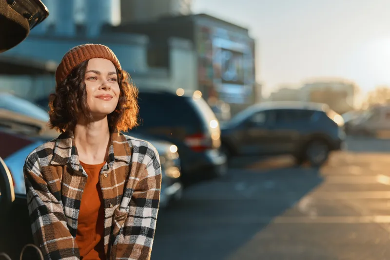 Woman smile in urban parking at golden hour glow, candid lifestyle portrait with beanie and plaid shirt, authenticity and mindful living captured in warm emotional storytelling.