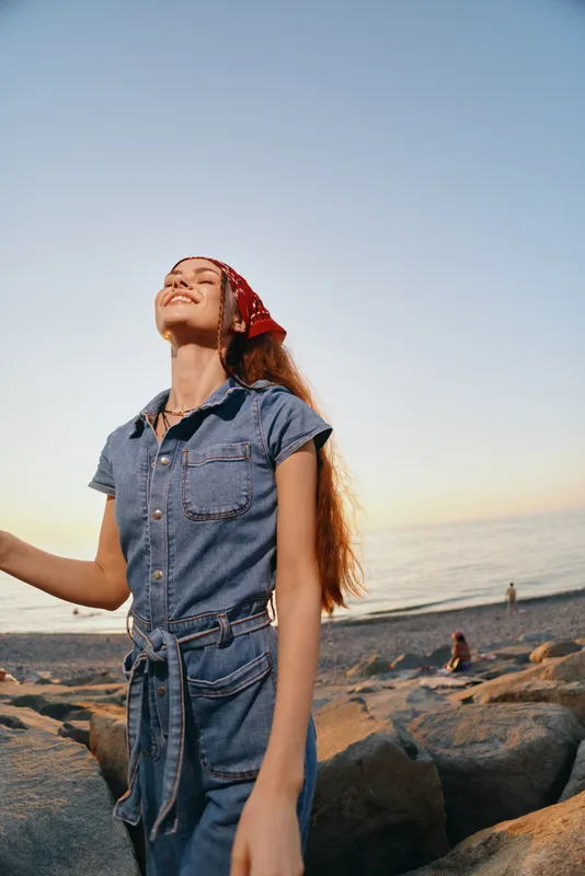 Lifestyle woman in modern boho-western denim street style enjoying sunset by the beach with film color tones and relaxed joyful expression.