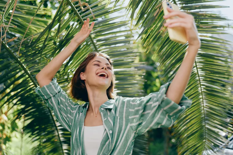 A joyful young woman takes a selfie beneath tropical palm leaves, basking in bright sunshine and carefree vacation vibes.