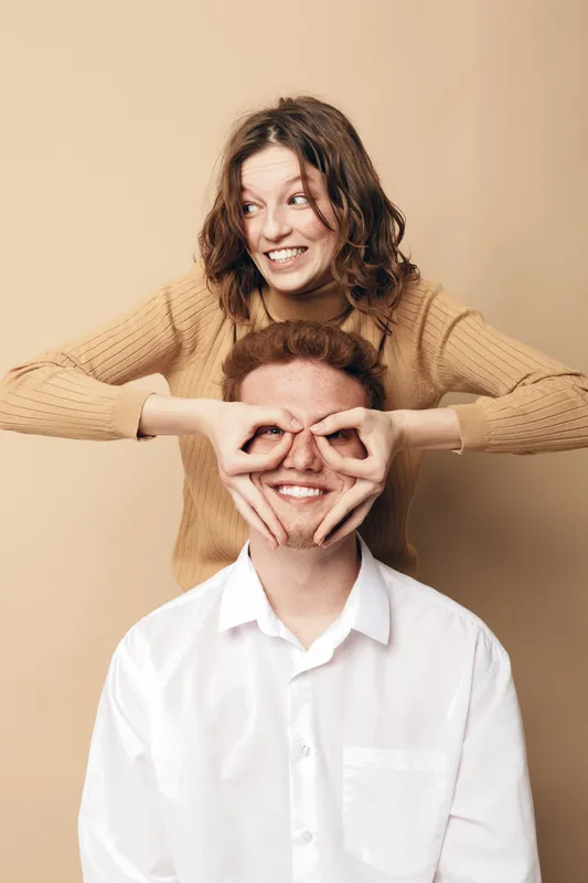Happy young woman in beige sweater playfully covers eyes of smiling man in white shirt. Studio portrait on beige background. Concept of friendship, fun, and joyful moments together.