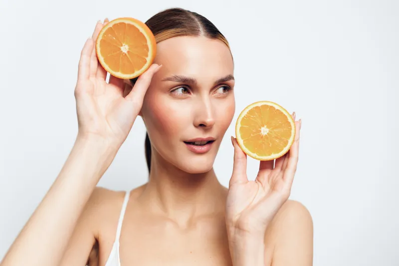 Woman beauty skincare concept, citrus orange halves held near eyes, freshness mood, studio light on white background, calm smile, gentle skincare routine, natural glow, minimal portrait