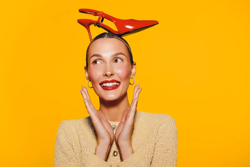 Smiling woman with red lipstick and gold earrings wears red high heel shoes on her head against bright yellow background. Concept of fashion, fun, creativity, and playful style.