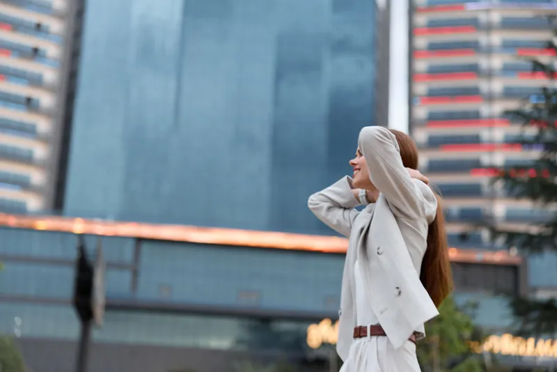 Young woman in beige suit enjoying city life with high-rise buildings background, joyful expression, modern business fashion concept