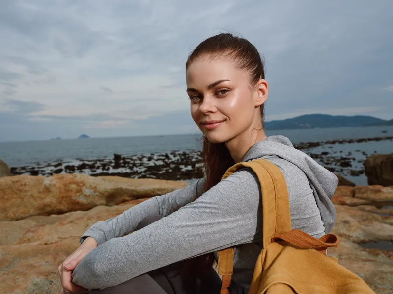 Smiling young woman with backpack on rocky shore, enjoying nature, outdoors fashion, serene seascape, natural beauty, casual style, spring vibe