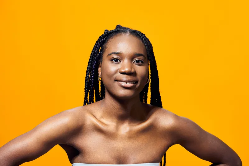 Confident young Black woman with braided hair smiling against a vibrant yellow background, showcasing joy and positivity in a fashion forward, seasonal portrait