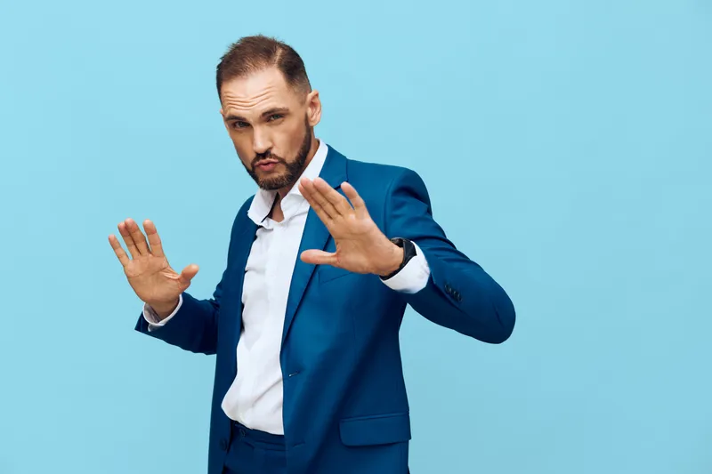 A professional man in a blue suit stands against a solid colored background, projecting confidence and caution as he raises hands in a stopping gesture.