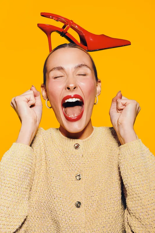 Excited woman with red lipstick and closed eyes holds fists near face while balancing red high heel shoe on head. Bright yellow background creates energetic and playful mood.