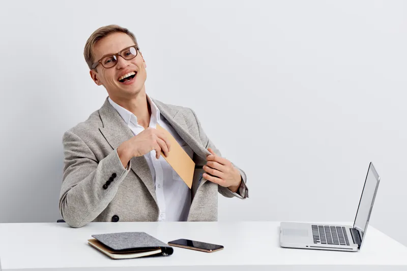 smiling young man in gray blazer putting tablet in jacket pocket sitting at desk with laptop smartphone and notepad digital nomad remote work concept