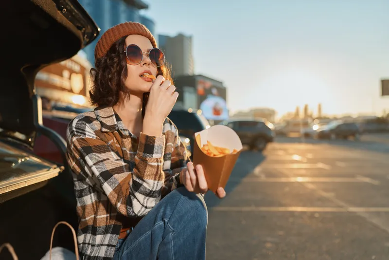 Woman eats fries from a car trunk in a parking lot at sunset, wearing sunglasses and a beanie in an urban scene. Authenticity, candid lifestyle and golden hour glow show mindful living.