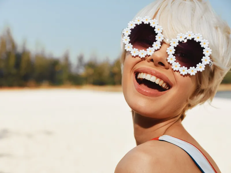 Joyful woman on a sunny beach wearing flowered sunglasses and a bright smile, capturing a carefree summer moment with warm light, vibrant energy, and relaxed style.
