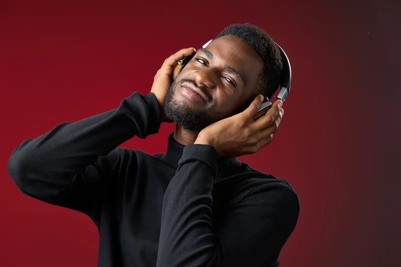 A young African man wearing headphones, enjoying music with a joyful expression against a deep red background, showcasing contemporary style and emotion in leisure
