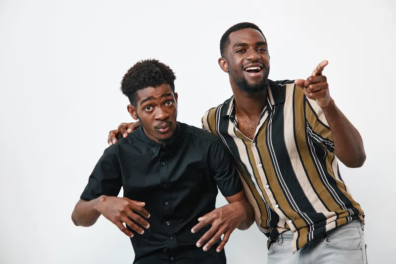 Two young black men with dark skin posing together in casual clothing on white background. One man wears a striped shirt and points forward while smiling, the other in black shirt looks curious.