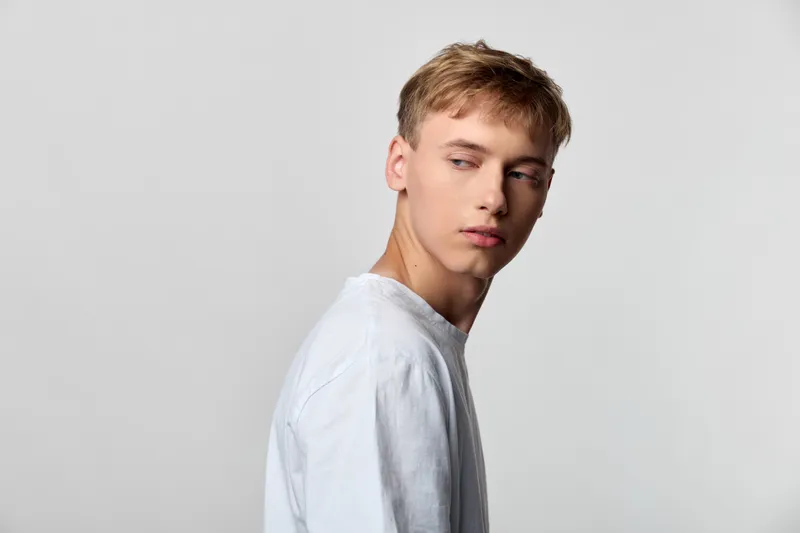 Young man in a plain white shirt turns his head to the side, a calm contemplative expression against a minimal studio backdrop, suitable for fashion, lifestyle, beauty or editorial concepts