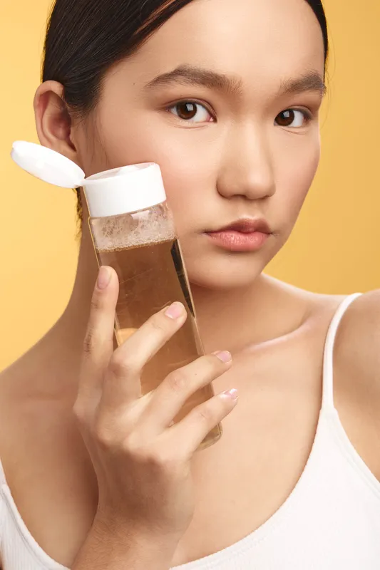 asian woman holds a skincare bottle close to her face in a bright minimal studio, calm expression, soft lighting and clean background, showcasing beauty routine and product focus.