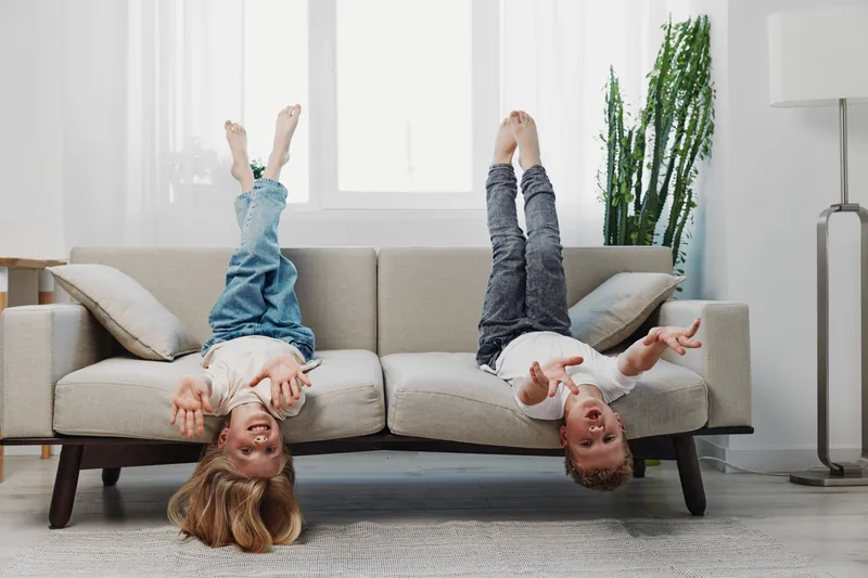 Two cheerful children hang upside down on a beige sofa in a bright living room, enjoying playful moments and carefree fun at home.