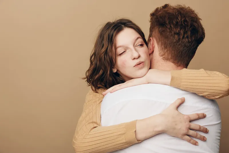 Young woman with brown hair hugs man wearing white shirt and beige sweater. Intimate moment of affection and comfort on neutral background. Concept of love and emotional support.