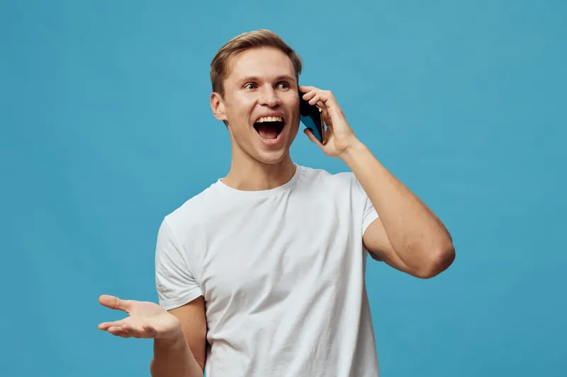 joyful man talking on mobile phone excited happiness cheerful expression casual white t-shirt blue background studio portrait. People lifestyle concept