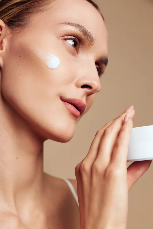 Woman closeup applying white face cream on her cheek, holding a small jar. Soft studio lighting on beige background. Skincare, selfcare, wellness routine, calm mood.