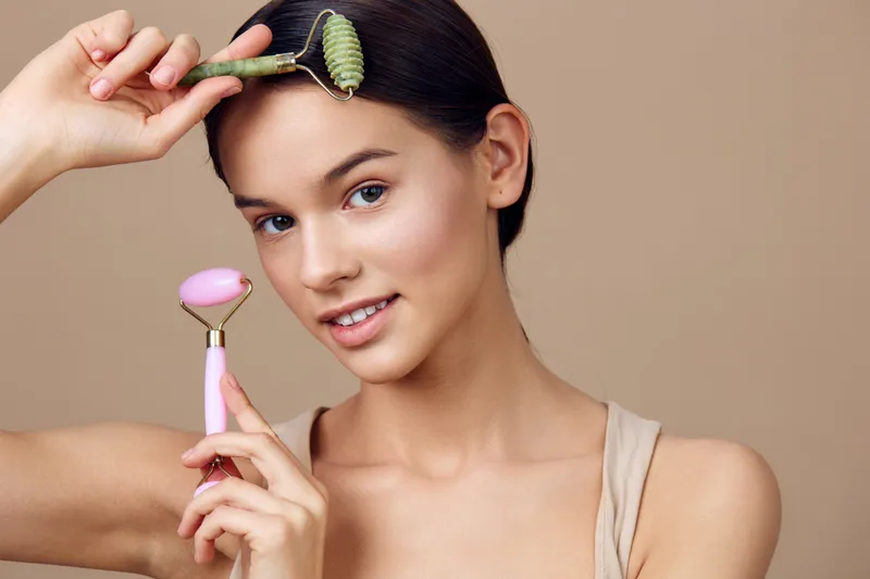 A young woman applying skincare rollers, showcasing a natural beauty routine with jade and rose quartz tools, emphasizing glowing skin and self care
