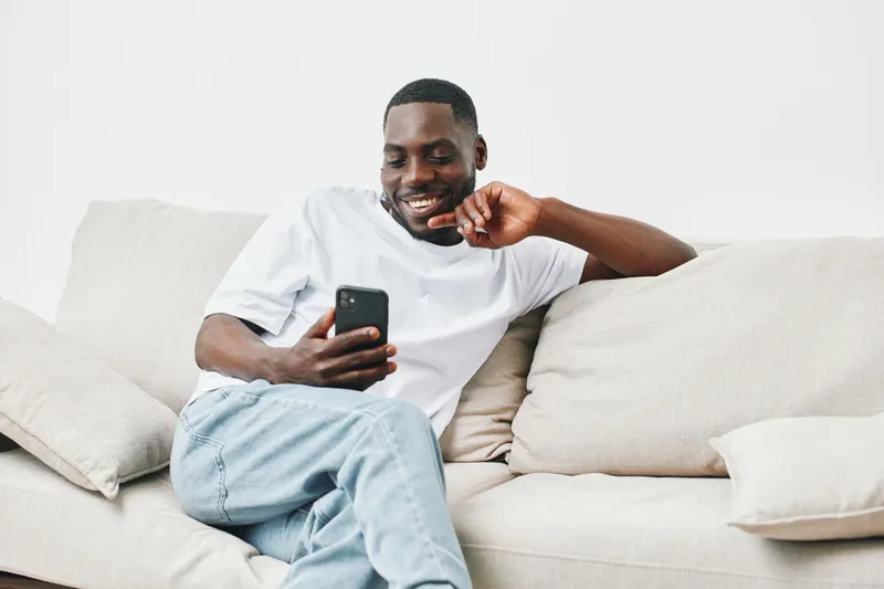 A smiling Black man in casual attire enjoying his smartphone on a comfortable sofa, conveying relaxation and modern lifestyle