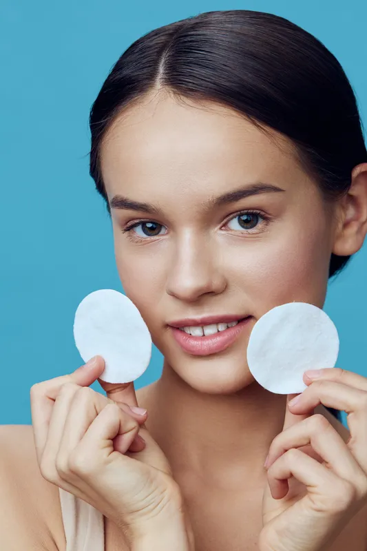 Young woman demonstrating skincare routine with cotton pads on blue background, highlighting healthy skin and beauty concept