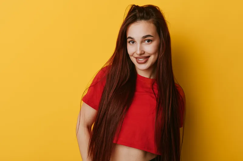 Young woman with long hair wearing red crop top smiling and posing against yellow background. Portrait of happy female with casual style and confident expression in studio.