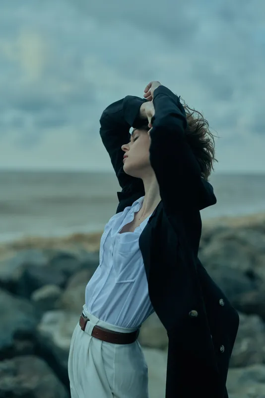 Windy coast fashion pose, woman lifts hair, wearing a white shirt and dark jacket, belt, standing with confidence against the sea horizon and rugged rocks, moody coastal scene