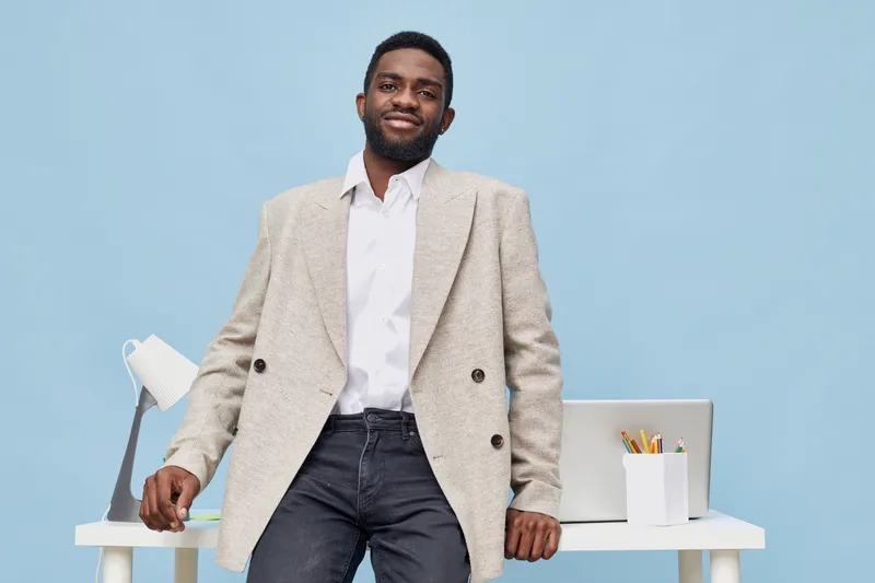 Confident smiling man standing in modern office with laptop and stationery on desk, wearing casual blazer and white shirt, light blue background, business casual style, professional atmosphere.