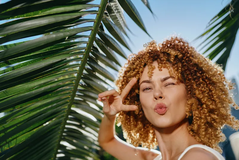 young woman with curly hair making peace sign near palm leaves under bright sunlight, summer outdoor portrait with natural green background